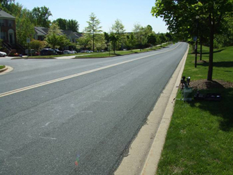 Photo shows the same street looking to the right -- it is straight for about 2 block-lengths.  On the left is an entrance into a housing development, on the right are trees and grass.